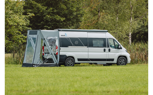 Witte camper met een grijs zijluifel bevestigd aan de zijkant op een grasveld, bomen en riet op de achtergrond