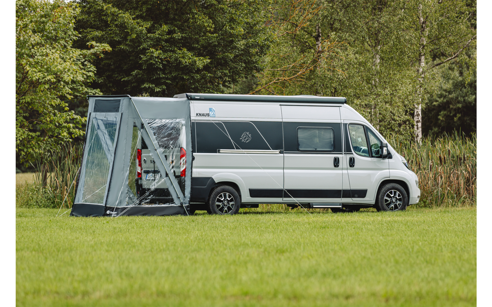 Witte camper met een grijs zijluifel bevestigd aan de zijkant op een grasveld, bomen en riet op de achtergrond