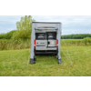 Achterkant van een witte camperbus met uitgevouwen luifel en open voortent op een grasveld, bomen en bewolkte lucht op de achtergrond