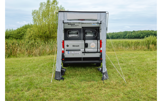 Achterkant van een witte camperbus met uitgevouwen luifel en open voortent op een grasveld, bomen en bewolkte lucht op de achtergrond
