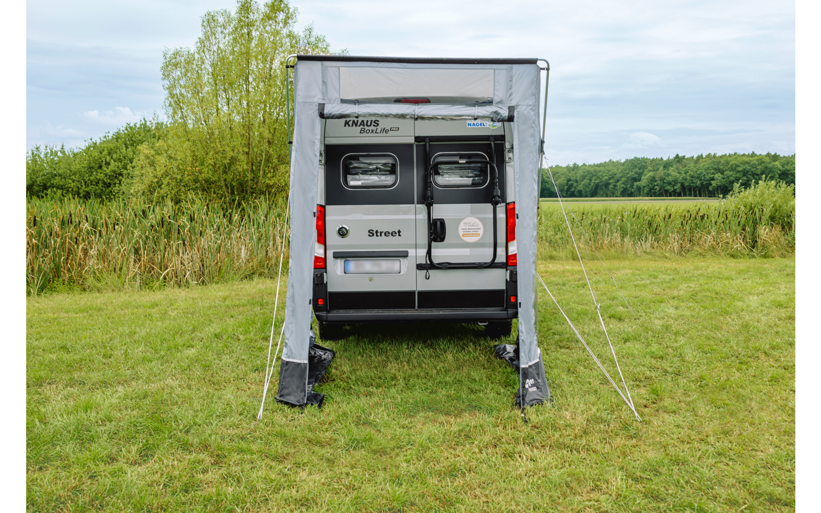 Achterkant van een witte camperbus met uitgevouwen luifel en open voortent op een grasveld, bomen en bewolkte lucht op de achtergrond