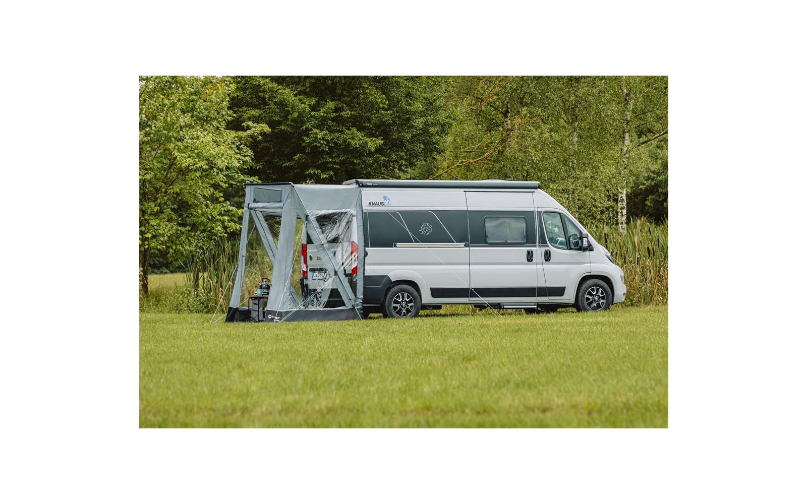 Weißer Camper mit grauem Vorzelt auf grüner Wiese vor herbstlichem Wald