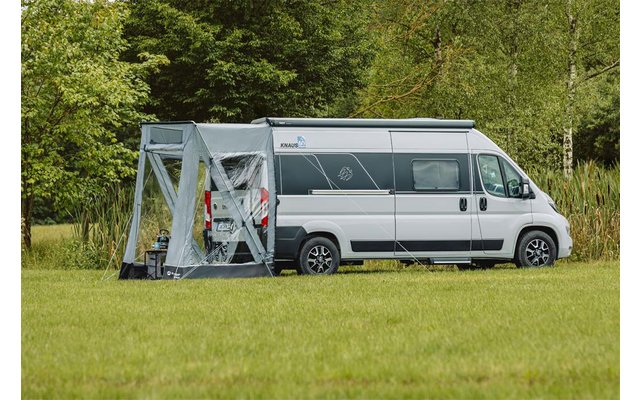 Weißer Camper mit grauem Vorzelt auf grüner Wiese vor herbstlichem Wald