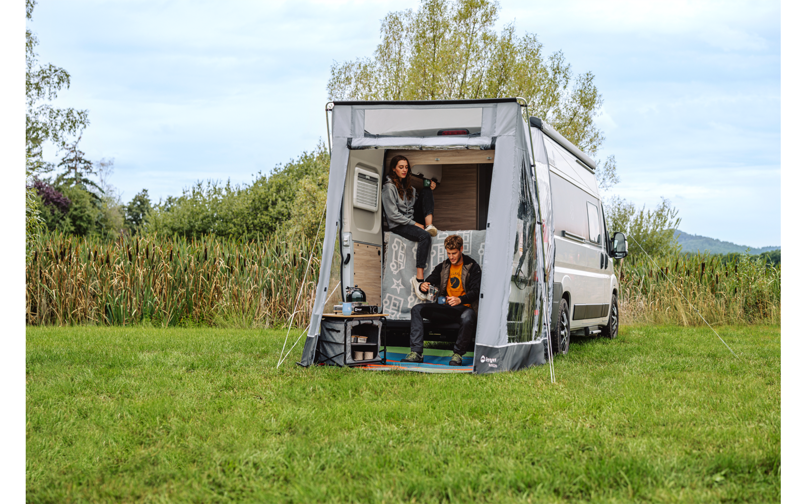Weißer Transporter mit geöffnetem Heck und angehängtem Vorzelt; zwei Personen, eine steht im Heck, eine sitzt an der Hecköffnung; Wiese, Schilf, Bäume und bewölkter Himmel