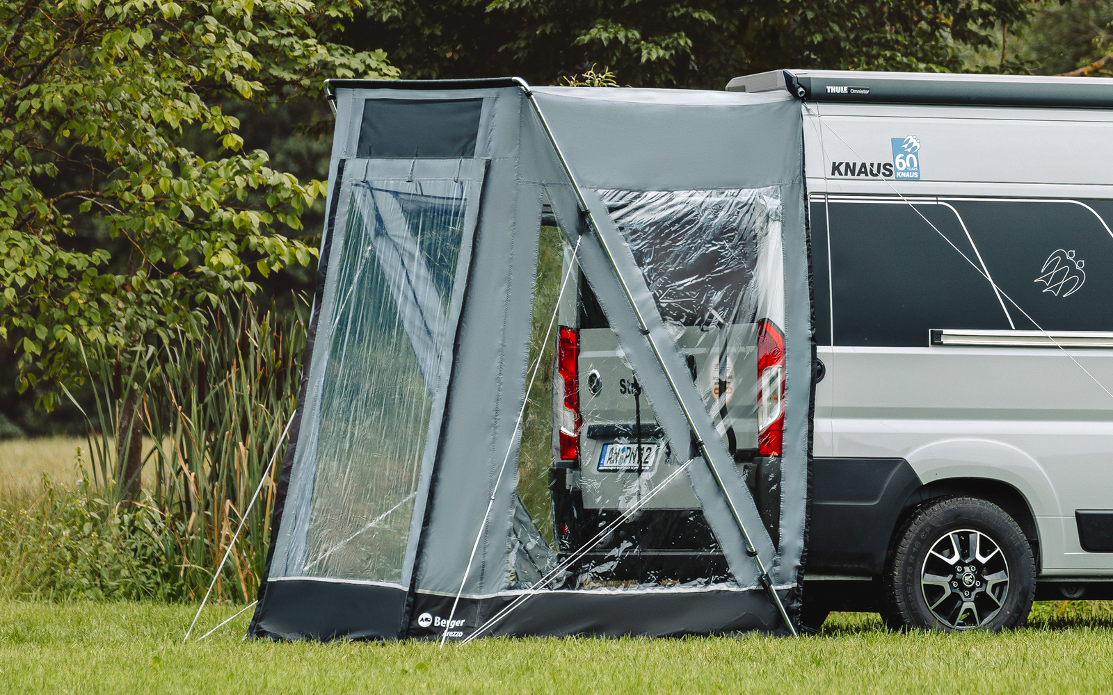 Weißes Wohnmobil mit grauem Vorzelt an der Seite auf einer Wiese, im Hintergrund Bäume und Schilf