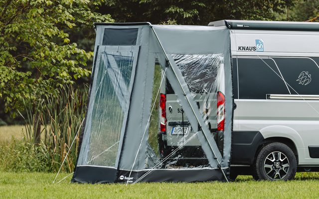 Weißes Wohnmobil mit grauem Vorzelt an der Seite auf einer Wiese, im Hintergrund Bäume und Schilf