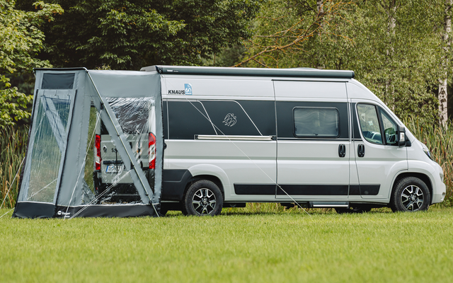 Autocaravana blanca con un toldo gris unido al lateral sobre un césped, árboles y juncos al fondo