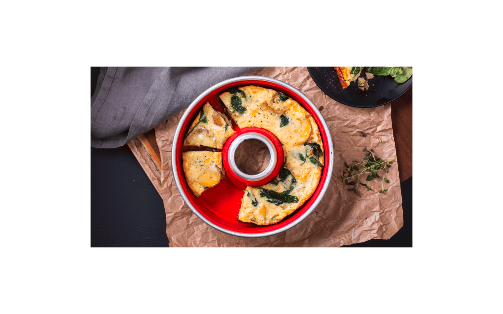 Round red baking pan with golden-brown, pie-slice portions containing green leaf pieces; on crumpled brown paper, gray cloth at left, black plate with one slice at top right, herb sprig at right