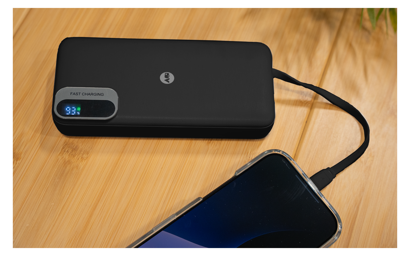 Schwarze Powerbank mit digitaler Anzeige "93%" und Aufschrift "FAST CHARGING" auf einer Holzoberfläche, per Kabel mit einem Smartphone verbunden
