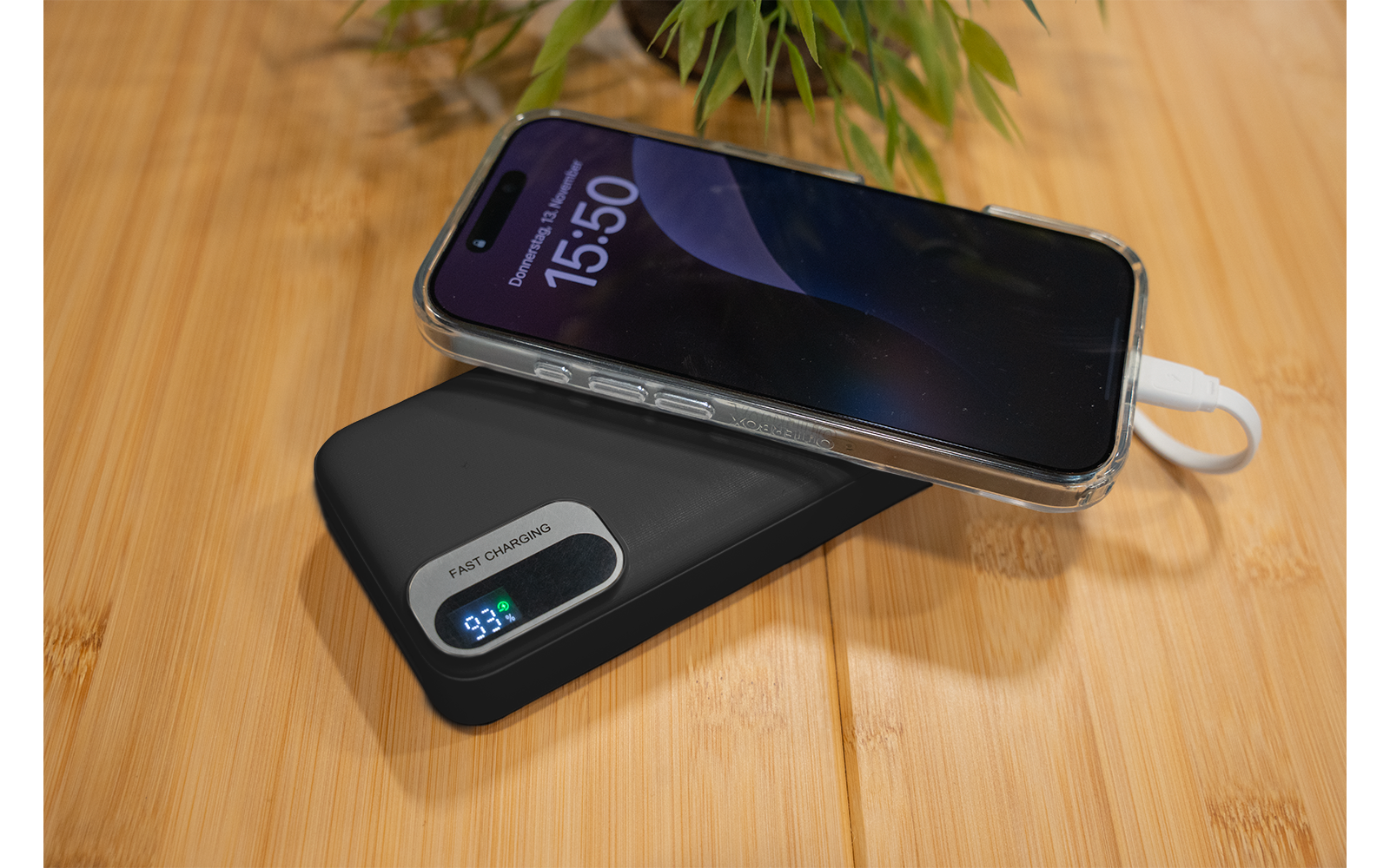 Teléfono inteligente sobre un power bank negro con pantalla digital '93%', cable de carga blanco visible, planta verde en maceta al fondo, superficie de madera