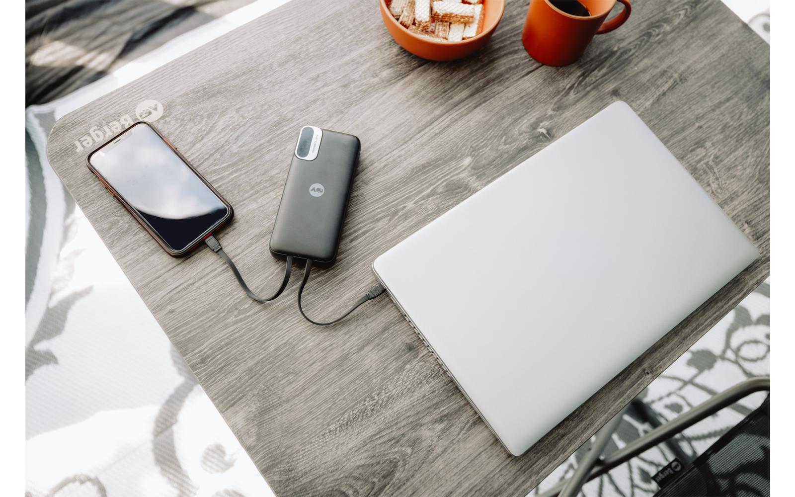 Geschlossener silberner Laptop auf einem Holztisch, ein Smartphone ist per Kabel mit einer Powerbank verbunden, eine Tasse und eine Schale mit kleinen rechteckigen Snacks