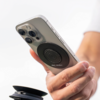 Hand hält ein Smartphone mit drei Kameralinsen und einem runden schwarzen Halter auf der Rückseite; unten links ein zusammengeklappter Ständer; weißer Hintergrund