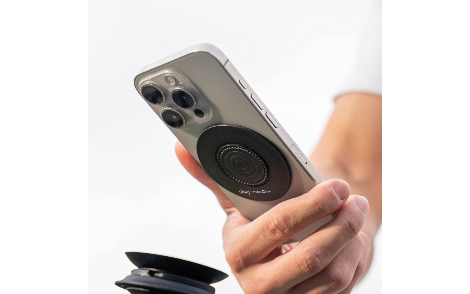 Hand hält ein Smartphone mit drei Kameralinsen und einem runden schwarzen Halter auf der Rückseite; unten links ein zusammengeklappter Ständer; weißer Hintergrund