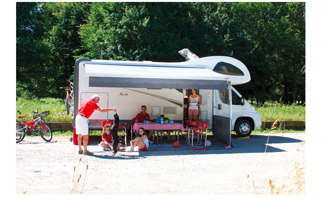 Wohnmobil steht auf einem Parkplatz, davor sitzen Personen an einem Tisch unter Sonnenschirm