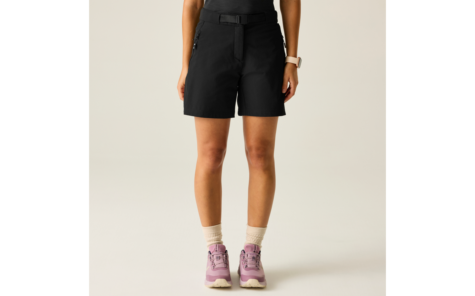 Persona desde las caderas hasta los pies; pantalones cortos negros, calcetines beige hasta el tobillo, zapatillas rosas; muñeca izquierda con reloj inteligente blanco; fondo liso y claro