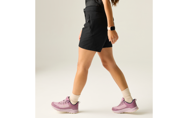 Parte inferior del cuerpo de una persona en posición de paso, falda corta negra, top negro, zapatillas rosas, calcetines cortos claros, reloj inteligente en la muñeca, fondo liso y claro