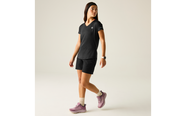 Persona con camiseta negra de cuello en V con pequeño logo y pantalones cortos negros, calcetines claros, zapatillas deportivas rosas, cinta en la cabeza y reloj en la muñeca, de pie ligeramente de perfil sobre fondo liso y claro