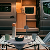 Offener Campingbus mit sichtbarem Holzinnenausbau; davor Klapptisch mit Thermoskanne, Schale mit Keksen, zwei Tassen und kleiner Lampe