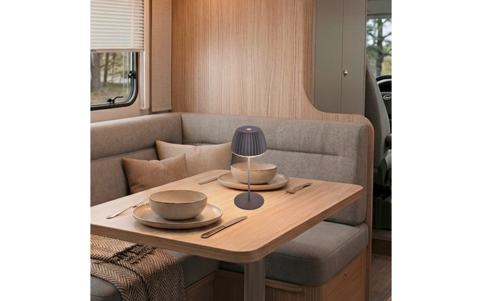 Holztisch mit kleiner Lampe in der Mitte, zwei gedeckte Plätze mit Schalen und Tellern, Besteck, graue Polsterbank mit Kissen, Fenster mit Jalousie, Lenkrad am rechten Bildrand sichtbar