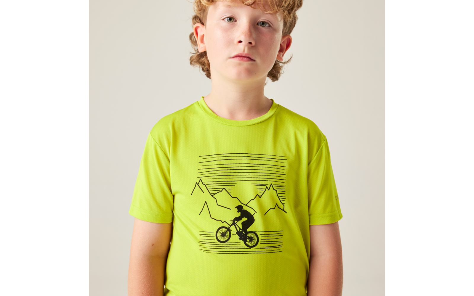 Kind mit lockigem blondem Haar, Oberkörper sichtbar, trägt leuchtgrünes T-Shirt mit schwarzem Aufdruck eines Mountainbikers vor einer Bergsilhouette, Blick nach vorn