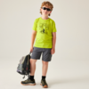 Junge mit lockigem Haar und Sonnenbrille, neongrünes T‑Shirt mit Fahrradmotiv, graue Shorts und Wanderschuhe, hält einen Rucksack, steht vor hellem Hintergrund