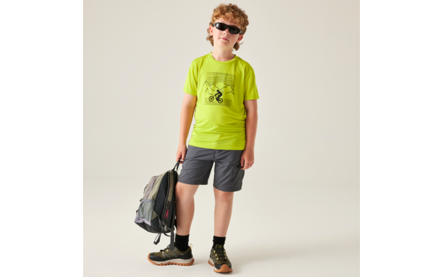 Junge mit lockigem Haar und Sonnenbrille, neongrünes T‑Shirt mit Fahrradmotiv, graue Shorts und Wanderschuhe, hält einen Rucksack, steht vor hellem Hintergrund