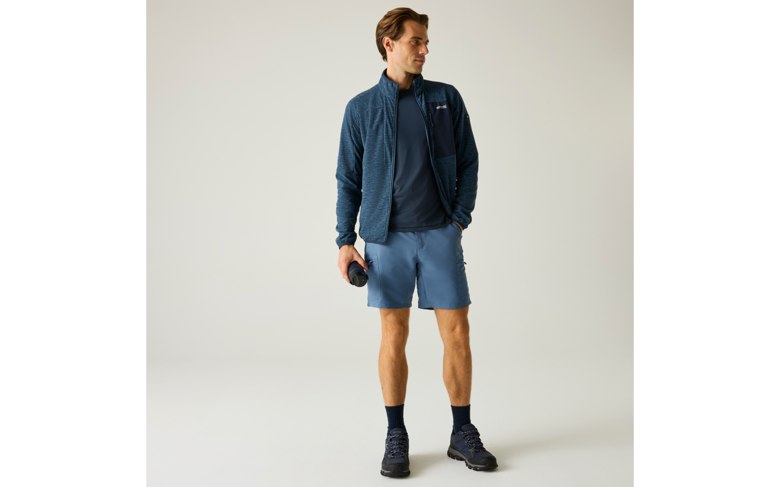 Homme debout devant un fond clair, porte une veste zippée bleu foncé, un T‑shirt sombre, un short bleu clair, des chaussettes foncées et des chaussures de randonnée, tient une gourde dans la main droite, regarde vers la gauche
