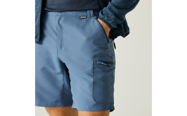 Bas du torse et cuisses d'une personne, short bleu avec poche zippée sur la cuisse, main droite dans la poche, pull bleu foncé visible