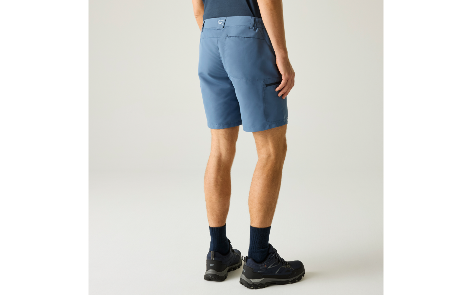 Personne vue de dos, portant un short bleu mi‑cuisse avec une poche latérale, chaussures sombres à lacets, chaussettes sombres aux chevilles, fond clair