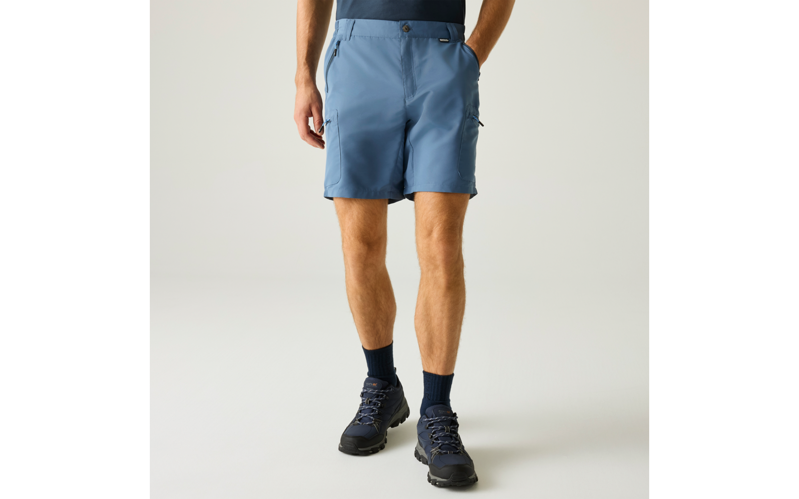 Bas du corps d'une personne portant un short bleu clair, une main dans la poche, chaussettes foncées, chaussures de randonnée foncées, fond gris clair