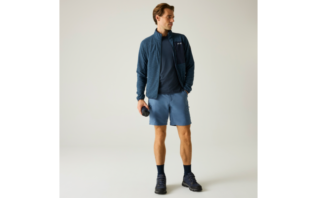 Homme debout devant un fond clair, porte une veste zippée bleu foncé, un T‑shirt sombre, un short bleu clair, des chaussettes foncées et des chaussures de randonnée, tient une gourde dans la main droite, regarde vers la gauche