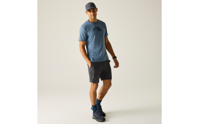 Person steht in blauem T-Shirt mit Logo, dunklen Shorts, blauer Kappe, blauen Socken und Wanderschuhen vor hellem Hintergrund