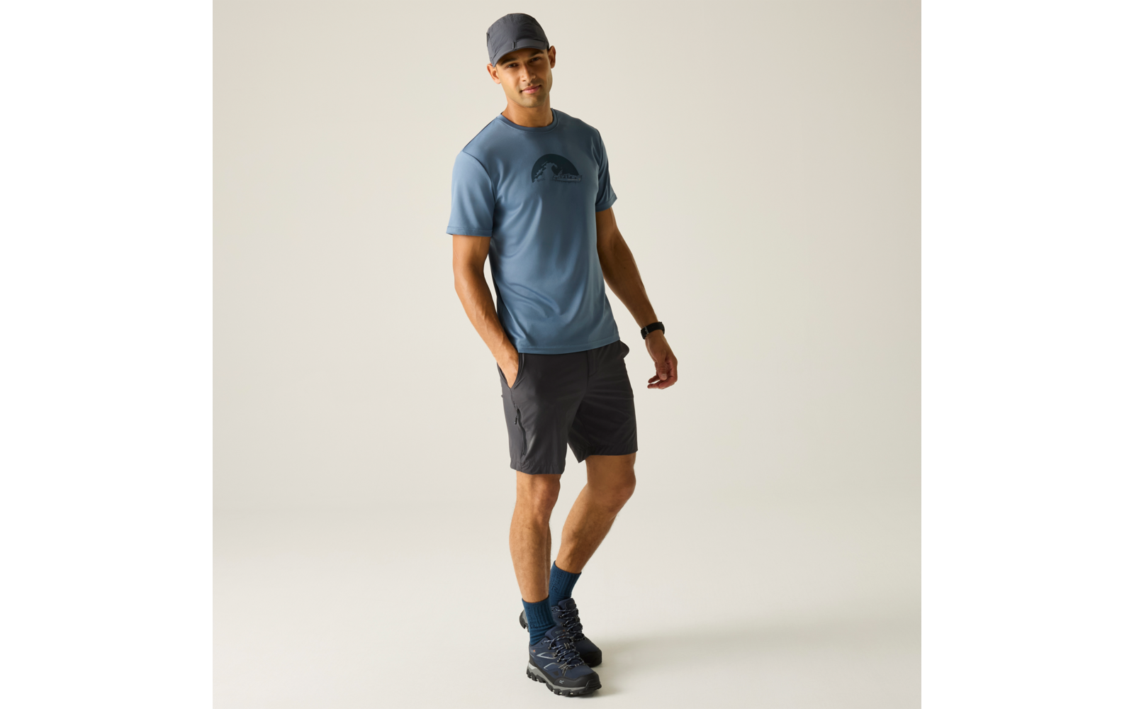 Person steht in blauem T-Shirt mit Logo, dunklen Shorts, blauer Kappe, blauen Socken und Wanderschuhen vor hellem Hintergrund