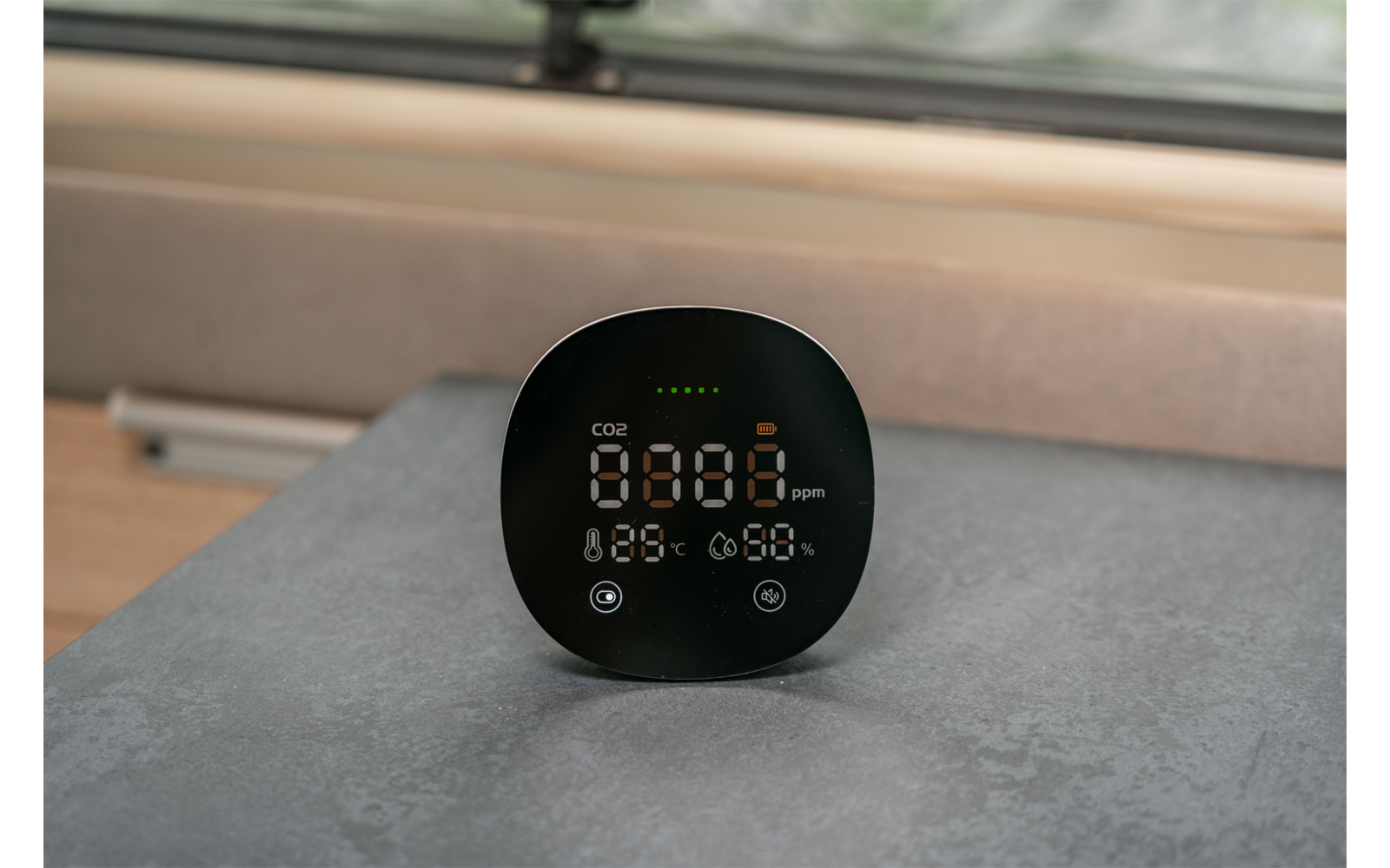 Rundes schwarzes Gerät mit digitaler Anzeige (Ziffern und Symbole), steht auf einer grauen Oberfläche vor einem Fenster