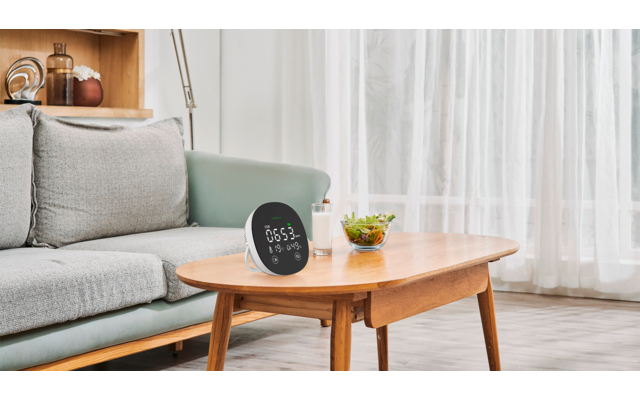 Graues Sofa, hölzerner Couchtisch mit rundem digitalen Display, auf dem '05:53' steht, kleine Schale mit grünen Blättern, Stehlampe, Regal mit Gläsern und Büchern, weiße transparente Vorhänge