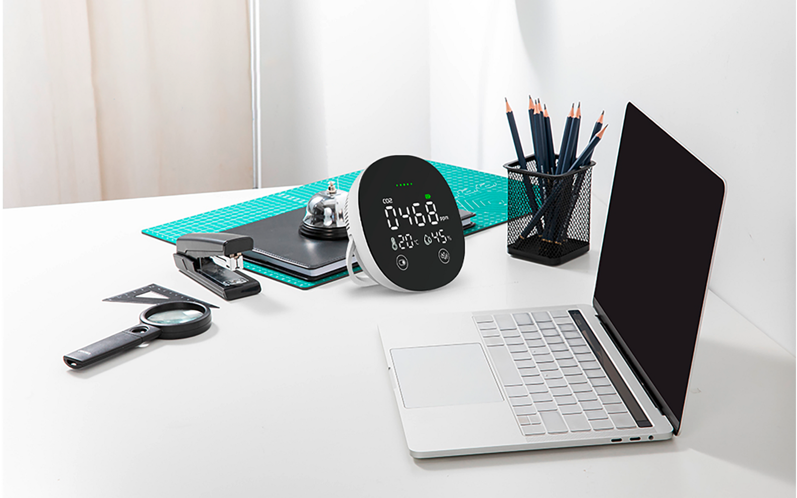 Weißer Schreibtisch mit offenem silbernem Laptop (schwarzer Bildschirm), rundes digitales Gerät mit Display, grüne Schneidematte, schwarzer Stiftehalter mit Bleistiften, Tacker, Lupe und silberne Glocke auf einem Notizbuch