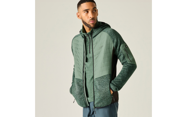 Mann mit kurzem Bart trägt grüne Kapuzenjacke mit Reißverschluss, Hände in den Taschen, blickt nach rechts vor hellem einfarbigem Hintergrund