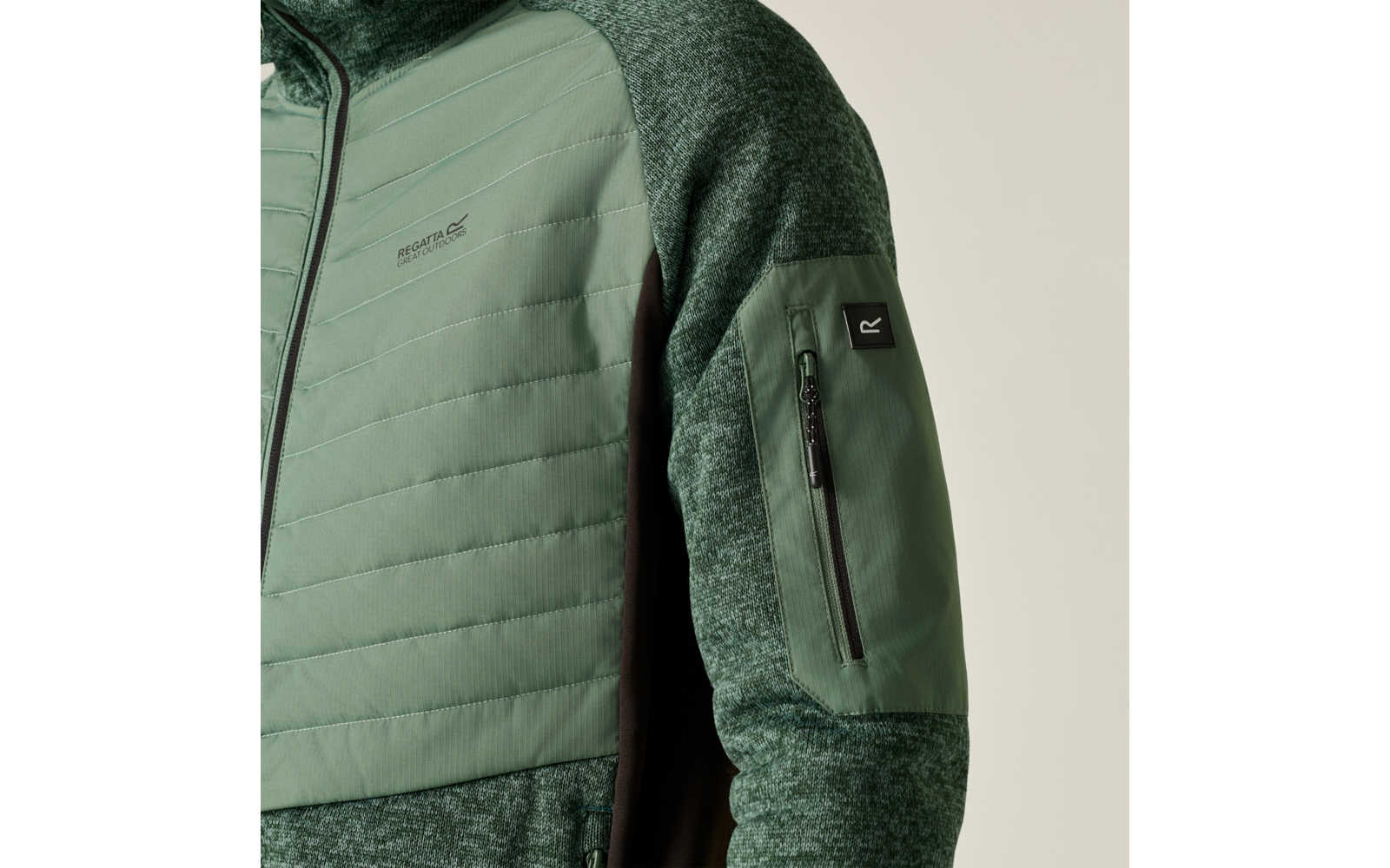 Nahaufnahme einer grünen Jacke mit gesteppter Vorderpartie, meliertem Strickärmel, Reißverschlusstasche am Oberarm und schwarzem Markenetikett