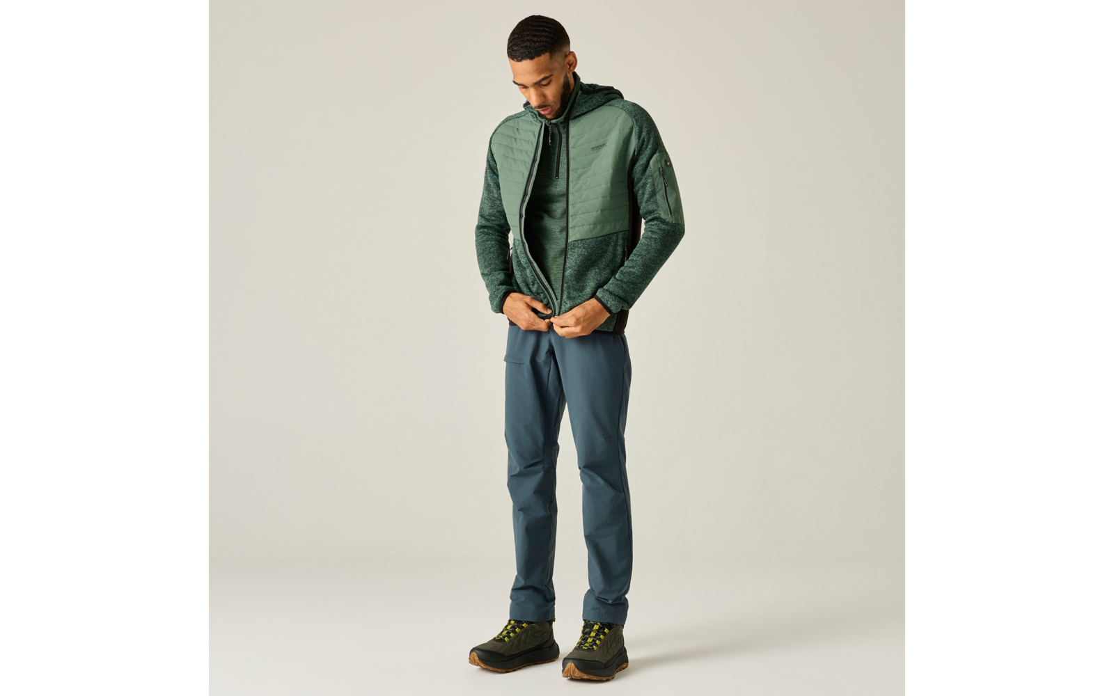 Mann ganzkörper stehend, schaut nach unten und schließt eine grüne Steppjacke; trägt blaue Hose und dunkle Wanderschuhe mit gelben Schnürsenkeln, heller einfarbiger Hintergrund