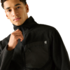 Person trägt schwarze Fleecejacke mit Reißverschluss und Brusttasche mit kleinem weißem Logo, eine Hand hält den Reißverschluss, beiger Hintergrund