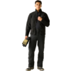 Person steht frontal, trägt schwarze Jacke, schwarze Hose und schwarze Schuhe, hält beige-schwarze Handschuhe in der linken Hand und fasst mit der rechten Hand den Jackenkragen. Heller einfarbiger Hintergrund