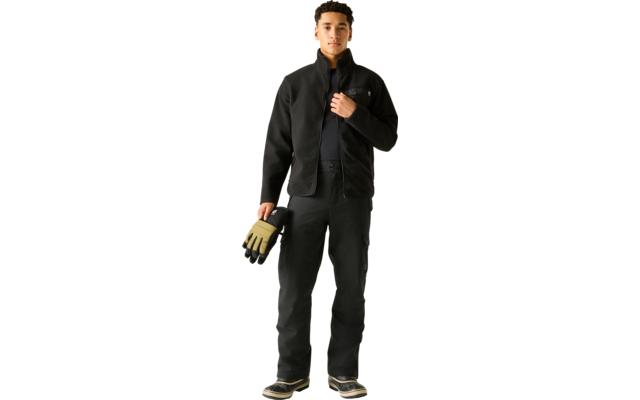 Person steht frontal, trägt schwarze Jacke, schwarze Hose und schwarze Schuhe, hält beige-schwarze Handschuhe in der linken Hand und fasst mit der rechten Hand den Jackenkragen. Heller einfarbiger Hintergrund
