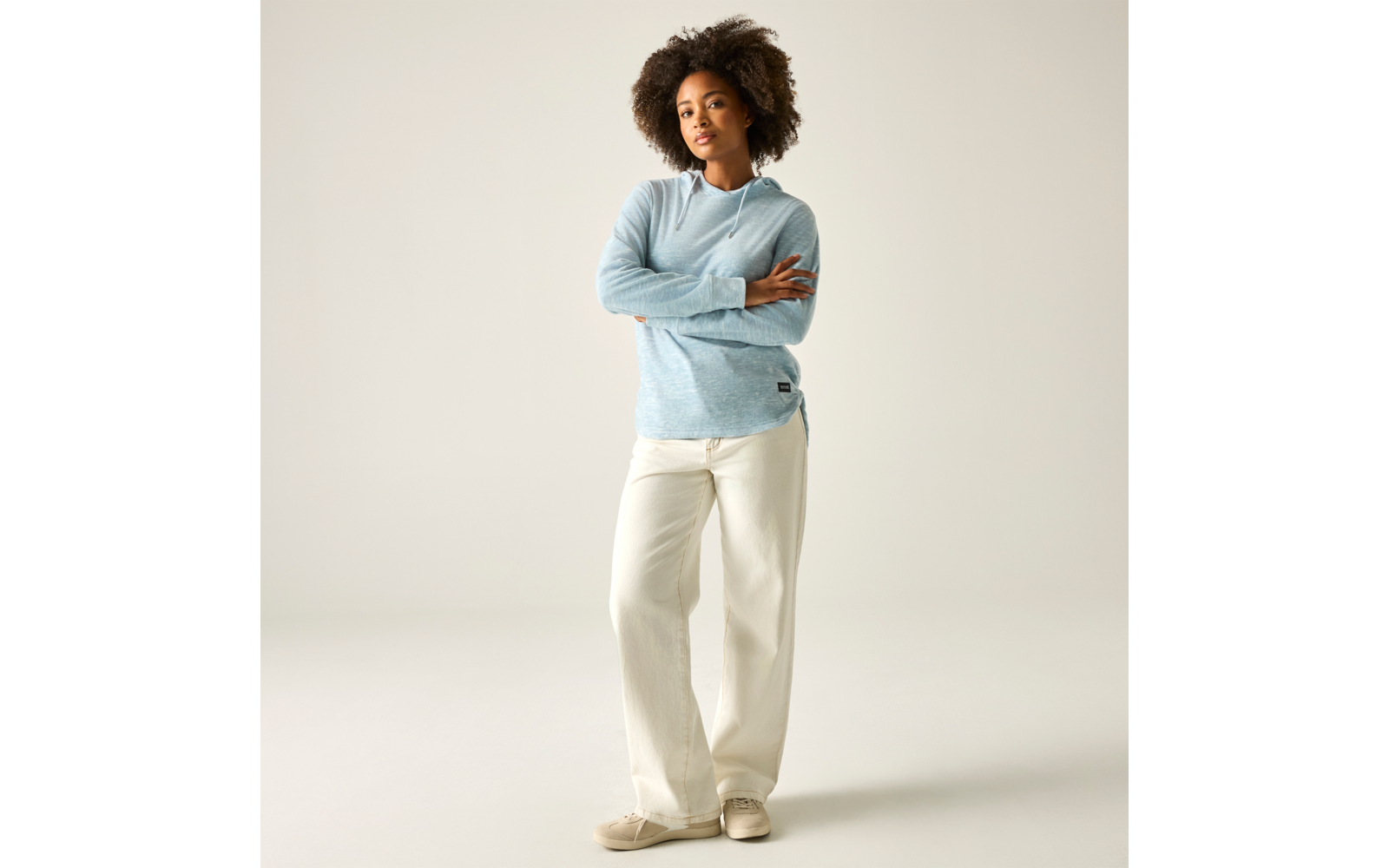 Person steht frontal, Arme verschränkt, trägt hellblauen Kapuzenpullover, weiße weite Hose und beige Schuhe vor hellem neutralem Hintergrund