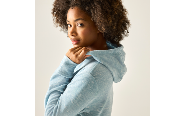 Person mit lockigem Haar trägt hellblauen Kapuzenpullover, blickt über die Schulter zur Kamera und hält mit einer Hand den Kapuzenrand; heller einfarbiger Hintergrund