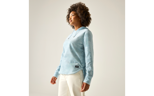 Frau mit lockigem Haar in hellblauem Hoodie und cremefarbenen Hosen, steht in Dreiviertelansicht und blickt nach oben, vor einfarbigem hellem Hintergrund