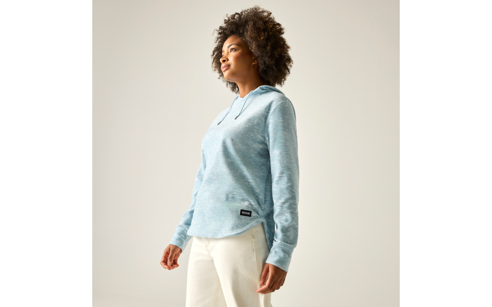 Frau mit lockigem Haar in hellblauem Hoodie und cremefarbenen Hosen, steht in Dreiviertelansicht und blickt nach oben, vor einfarbigem hellem Hintergrund