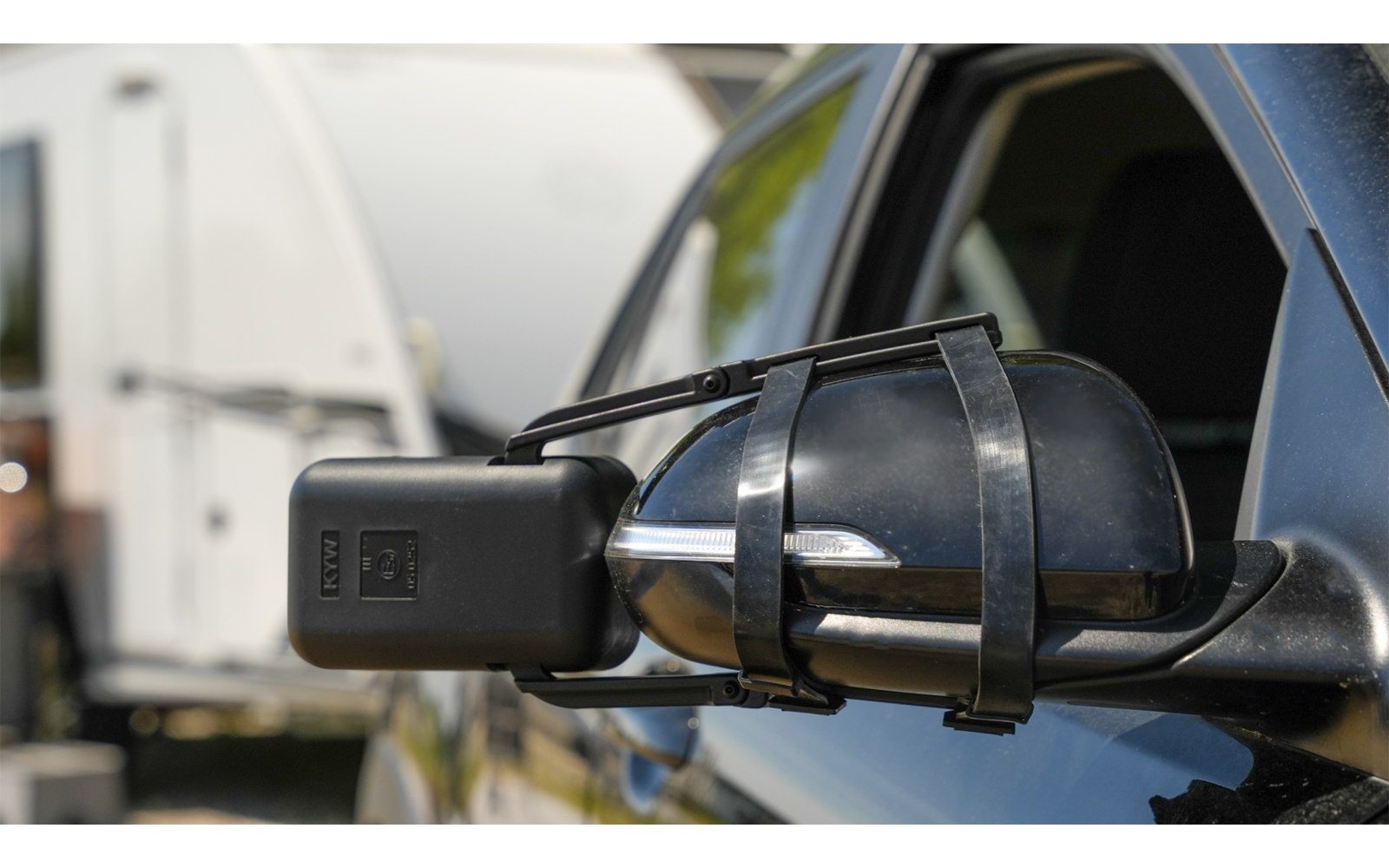 Seitenspiegel eines schwarzen Fahrzeugs mit einem daran befestigten Zusatzspiegel, im Hintergrund ein weißes Wohnmobil