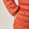 Nahaufnahme einer orangefarbenen Steppjacke (Ärmel und Oberkörper), rechte Hand in Tasche, weiße Aufschrift FEATHER FREE auf dem Ärmel, heller Hintergrund