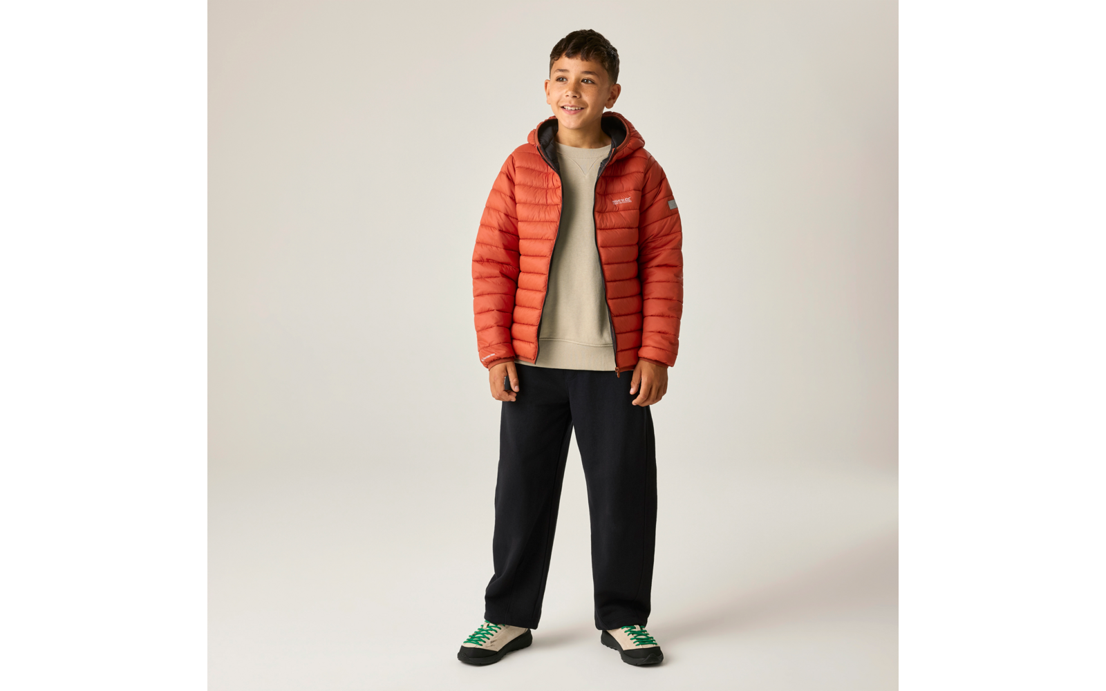 Junge steht vor neutralem Hintergrund, trägt orange Steppjacke, beigefarbenen Pullover, schwarze Hosen und weiße Sneakers mit grünen Schnürsenkeln