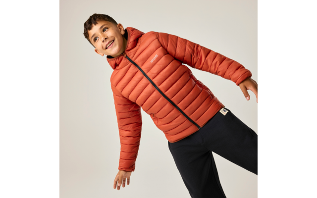 Junge in oranger Steppjacke mit Kapuze und schwarzer Hose, leicht nach rechts geneigt vor hellem Hintergrund